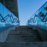 Concrete stairs with glass railings reflecting blue sky, captured from a low angle.
