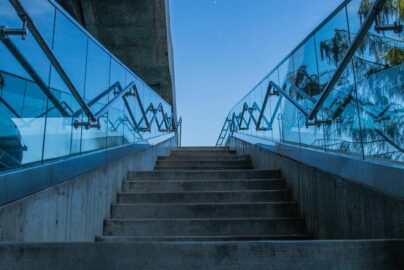 Concrete stairs with glass railings reflecting blue sky, captured from a low angle.