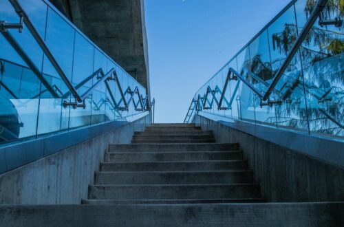 Concrete stairs with glass railings reflecting blue sky, captured from a low angle.