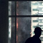 A silhouette inside observes a dramatic high-rise blaze, creating a tense and intense urban scene.