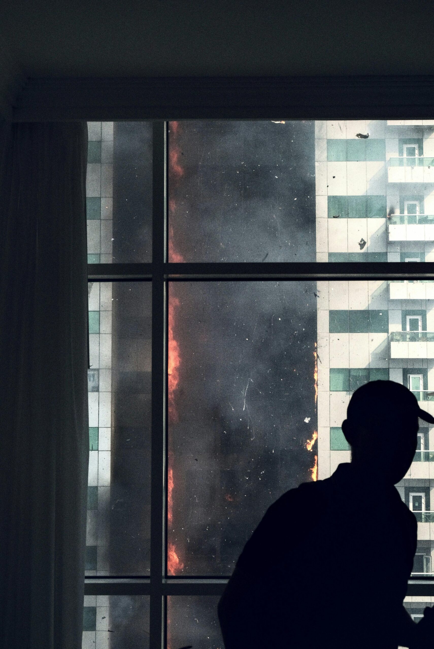A silhouette inside observes a dramatic high-rise blaze, creating a tense and intense urban scene.